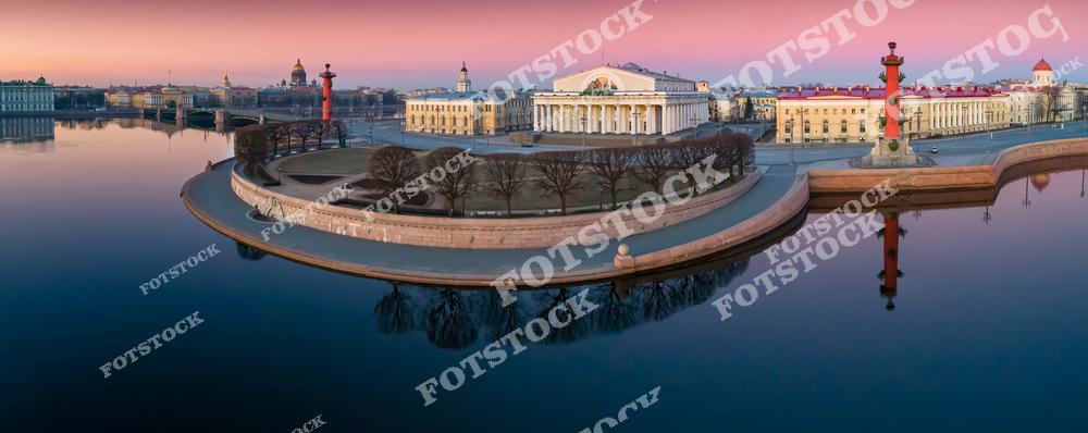 На фотографии изображена Стрелка Васильевского острова — одна из самых узнаваемых достопримечательностей Санкт-Петербурга. В центре композиции — здание Старой Биржи, построенное в стиле классицизма. По бокам — Ростральные колонны, служившие в XIX веке маяками для судов. Спокойные воды Невы отражают величественную архитектуру и розовое небо рассвета. Слева в глубине виднеются купола Исаакиевского собора и здания Адмиралтейства.